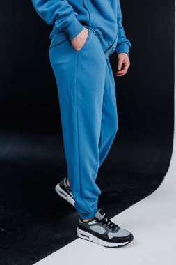 Bir insan şık siyah spor ayakkabılarıyla uyumlu rahat mavi bir eşofman giyiyor. Kıyafetleriyle sıradan ama modaya uygun bir ifade sergiliyor.