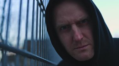 Koyu renk kapüşonlu bir adam yıpranmış bir çitin yanında duruyor, eşsiz, cesur bir şehir atmosferini somutlaştırıyor.