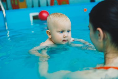 Bebek havuzda yüzüyor. Bir eğitmenin gözetiminde bebekler için yüzme havuzu. Havuzda çocuğu olan genç bir anne. Elinde bebekle suda eğlenen bir kadın.