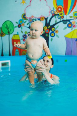 Bebek havuzda yüzüyor. Bir eğitmenin gözetiminde bebekler için yüzme havuzu. Havuzda çocuğu olan genç bir anne. Elinde bebekle suda eğlenen bir kadın.