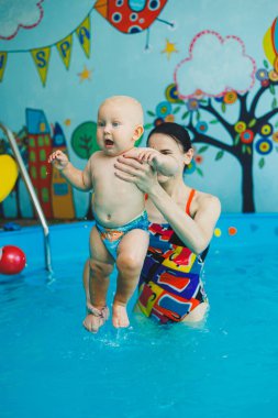 Bebek havuzda yüzüyor. Bir eğitmenin gözetiminde bebekler için yüzme havuzu. Havuzda çocuğu olan genç bir anne. Elinde bebekle suda eğlenen bir kadın.