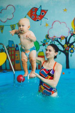 Bebek havuzda yüzüyor. Bir eğitmenin gözetiminde bebekler için yüzme havuzu. Havuzda çocuğu olan genç bir anne. Elinde bebekle suda eğlenen bir kadın.