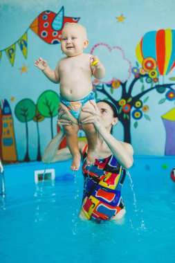 Bir eğitmenin gözetiminde yüzen bebekler için havuz. Havuzda çocuğu olan genç bir anne. Kadın kucağında bebek, suda eğleniyor.