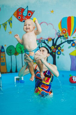 Bebek havuzda yüzüyor. Bir eğitmenin gözetiminde bebekler için yüzme havuzu. Havuzda çocuğu olan genç bir anne. Elinde bebekle suda eğlenen bir kadın.