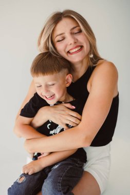 Yürek ısıtan bir sahnede, gülümseyen bir anne hem saf aşk hem de mutluluk saçan neşeli oğlunu sevgiyle kucaklar, basit, parlak bir stüdyo ortamında yakalanır.