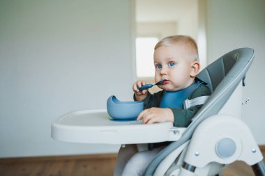 Aile hayatının özünü güzel bir şekilde somutlaştıran parlak ve minimalist bir odada, şirin bir bebek yüksek bir sandalyede oturur, kaşıkla çeşitli yiyecekleri neşeyle keşfeder.