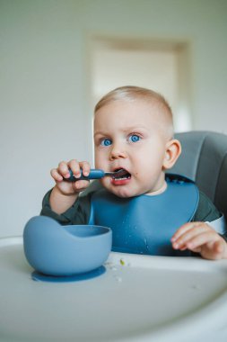 Yüksek sandalyedeki neşeli bir bebek, renkli bir kaşık ve kâseyle oynayan, parlak mavi gözleri olan ve odayı aydınlatan neşeli bir yüz ifadesi olan yemek vaktinden zevk alır.
