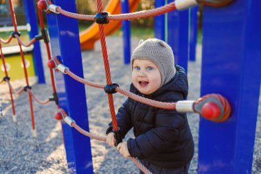 Neşeli genç bir çocuk neşeli bir oyun alanında oynuyor, halatlara tırmanıyor ve oyunlara katılıyor, sonbahar gökyüzünün altında sıcacık bir kış şapkası ve ceket giyiyor.