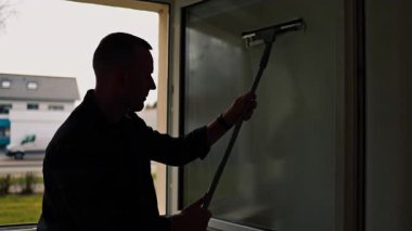 Pencere temizliği. Camları paspasla siliyorum. Bir adam evdeki bir paçavrayla pencereleri dikkatlice siler. Odada temizlik ve düzen var. Ev hanımı çalışıyor.