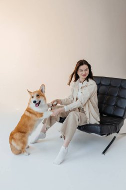 Neşeli genç bir kadın, sevgi dolu Corgisiyle neşe içinde etkileşime geçiyor. Evcil hayvanlarla sahipleri arasındaki samimi ve mutlu bağı vurguluyor.