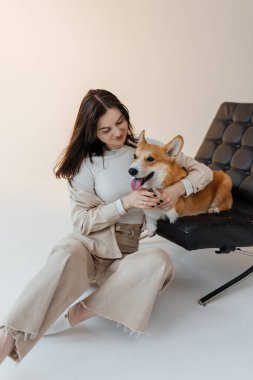 Minimalist bir stüdyoda, parlak bir gülüşü olan genç bir kadın neşeli Corgi 'sini kucaklıyor, samimi bir arkadaşlık ve şefkat anı yakalıyor.