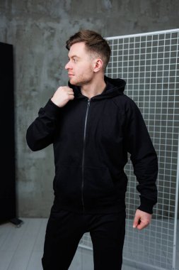 Siyah fermuarlı kapüşonlu şık bir genç adam ve eşgüdümlü pantolonlar şık ve çağdaş bir arkaplan önünde rahatça poz veriyor, havalı bir zarafet hissi yayıyor.