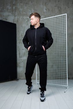 Gösterişli siyah atletizm kıyafetleri giymiş şık ve kendinden emin genç bir adam spor, moda veya yaşam tarzı temalarını yakalamak için mükemmel bir şekilde modern bir stüdyo ortamında poz veriyor.