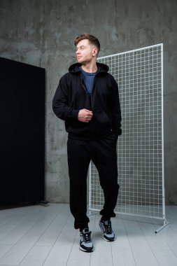 Tamamen siyah spor kıyafetleri giymiş şık bir genç adam endüstriyel bir ortamda poz veriyor, modern stili ve atletik moda seçimini etkili bir şekilde sergiliyor.