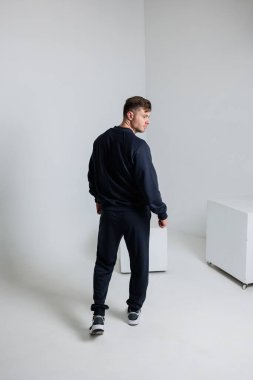 Siyah atletizm kıyafetleri giymiş genç bir adam sırtı dönük bir şekilde modern minimalist stüdyoda duruyor. Hem fitness hem de çağdaş moda trendlerini sergiliyor.