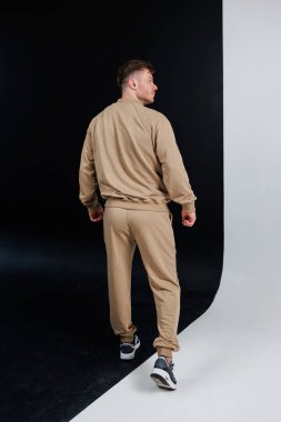 Bir erkek model, siyah ve beyaz zıt arkaplanlarla dolu bir stüdyoda güzel bir şekilde çekilen bej renkli spor kıyafetini güvenle sergiliyor.