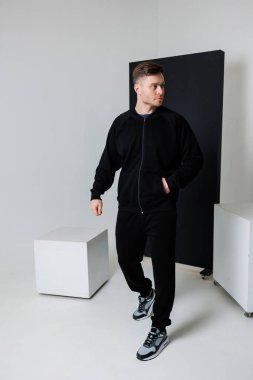 Kendine güvenen genç bir adam zarif siyah eşofmanlarıyla minimalist bir stüdyo arkaplanına karşı modern atletizm tarzını sergileyerek zarafetle yürür.