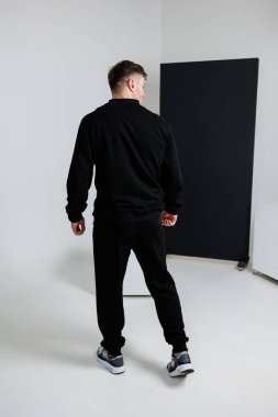 Gösterişli siyah eşofman giyen bir erkek model, sırtı kameraya dönük bir şekilde minimalist bir stüdyoda son moda spor kıyafetlerini sergiliyor.