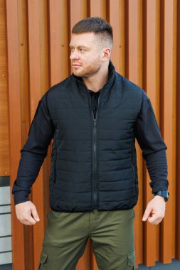 Kendine güvenen bir adam kendine güvenen bir siyah kabarık yelek ve şık bir kıyafet ile modern bir şehir arka planına karşı, en son moda eğilimlerini sergiliyor.