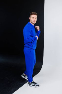 Kendine güvenen genç bir adam şık bir fotoşopla canlı bir mavi eşofman sergiliyor. Modern spor giyim estetiğini temiz ve minimalist bir arka plana karşı vurguluyor.
