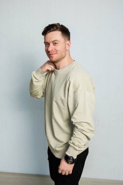 Kendinden emin genç bir adam yüzünde neşeli ve anlamlı bir ifadeyle poz veriyor, açık renkli uzun kollu bir gömlek giymiş, basit ama dokulu bir arka plana yerleştirilmiş.