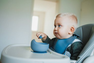 Bir buçuk yaşındaki bir çocuk yüksek bir sandalyeye oturur ve bağımsız olarak kaşıkla silikon tabaktan çorba içer. Çocukları beslemek için silikon kaplar. Çocuk kendi başına yemek yemeyi öğreniyor..