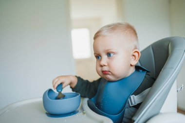 Gülümseyen çocuk sandalyede oturmuş çorba içiyor. Aç çocuk ağzına yemek koyuyor. Bebek 1 yaşında ve 3 aylık, tek başına yemek yiyor. Çocukları beslemek için silikon kaplar.