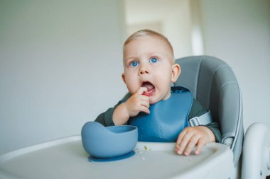 Mavi gözlü çocuk, elinde silikon bir önlükle sandalyede oturmuş, kendi başına yemek yiyor. Küçük çocuk kendi başına yemek yemeyi öğreniyor. Çocukları beslemek için güvenli aletler.