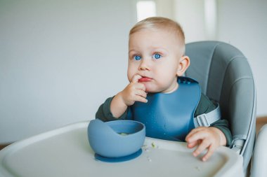 Mavi gözlü çocuk, elinde silikon bir önlükle sandalyede oturmuş, kendi başına yemek yiyor. Küçük çocuk kendi başına yemek yemeyi öğreniyor. Çocukları beslemek için güvenli aletler.