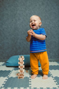 Evde çocuk odasında koşuşturan bir yaşında bir çocuk. Tişörtlü ve pantolonlu çocuk evde oyuncaklarla oynuyor. Çocuk kıyafetleri. Çocuğun evde kendi kendini geliştirmesi.