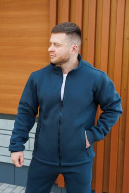 Şık bir donanma ceketi giymiş, kendine güvenen genç bir adam şık bir pantolona sahip, modern ahşap mimarinin çarpıcı arka planına karşı açık havada poz veriyor.