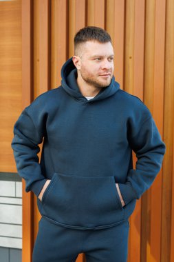 Tarz sahibi, koyu renk bir kapüşonlu giyen genç bir adam, açık havada, dikkat çekici ve modern şehir arkaplanının ortasında, hiç çaba harcamadan moda ve rahat bir moda anlayışı sergiliyor.