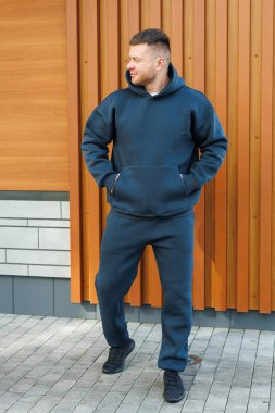 Genç bir adam, modaya uygun koyu gri kapüşonlu, rahat eşofman giyen, modern gündelik modanın özünü özetleyen, kırsal ahşap bir duvara karşı güvenle poz veriyor.