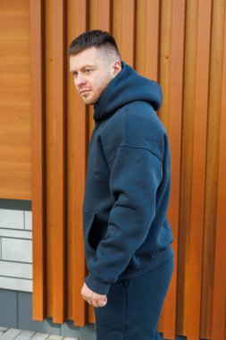 Modayı takip eden bir adam, büyük, koyu renk kapüşonlu bir kapüşonlunun içinde, güzel ve zıt ahşap dokulara karşı şık bir şekilde modern tarzını sergiliyor.