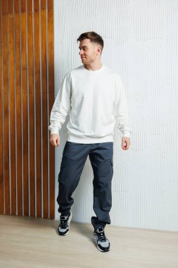 Bir erkek model modaya uygun beyaz bir sweatshirt ve koyu renkli bir kargo pantolonuyla poz veriyor, canlı bir şehir arka planına karşı modern sokak kıyafetleri somutlaştırıyor, çağdaş moda sergiliyor.