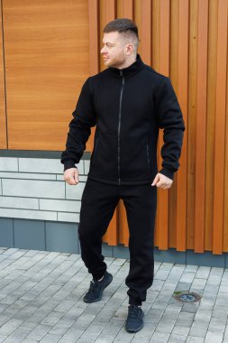 Gösterişli, siyah bir spor kıyafeti giymiş kendine güvenen bir adam dışarıda duruyor, modern tarz ve konforun modern bir karışımını çağdaş bir arka planda sergiliyor.