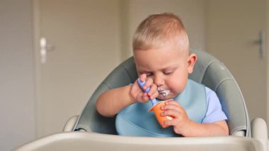Bir yaşındaki bir çocuk masaya oturur ve tek başına süt yoğurdu yer. Çocuk bebek yoğurdu yiyor. Çocuk lezzetli bir şekilde yoğurt yiyor ve yüzüne bulaştırıyor..