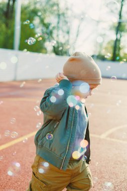 Küçük çocuk sabun köpüğüyle dışarıda koşuyor. Çocuk sıcak bir bahar gününde ceket ve şapkayla yürüyüşe çıkıyor..
