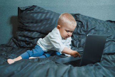 2 yaşında bir çocuk yatakta oturur ve dizüstü bilgisayardaki tuşlara basar. Çocuk bir dizüstü bilgisayarla oynuyor..