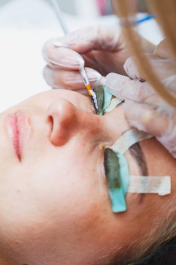 Bir kadının yüzünde kirpik kaplama prosedürü. Bir kozmetolog, kirpik uzatma işlemi sırasında bir kadının gözüne cımbızla uzun kirpikleri yapıştırır..