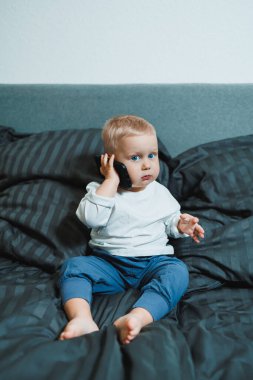 İki yaşında beyaz tişörtlü bir çocuk elinde bir telefonla yatağın üzerinde oturuyor. Modern küçük çocukların dijitalleştirilmesi. Küçük çocuk cep telefonu eğitimi alıyor..