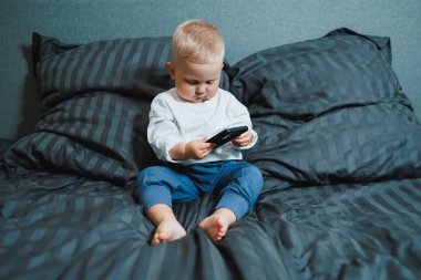 İki yaşında beyaz tişörtlü bir çocuk elinde bir telefonla yatağın üzerinde oturuyor. Modern küçük çocukların dijitalleştirilmesi. Küçük çocuk cep telefonu eğitimi alıyor..