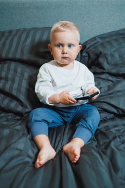 Neşeli küçük çocuk bütün gün yatakta oturup akıllı telefondan oyun oynuyor. Evde akıllı telefon tutarak yatağında oturan çocuğu yakından çek. Cep telefonuyla gülümseyen çocuk.