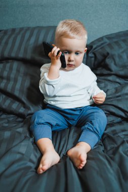 İki yaşında beyaz tişörtlü bir çocuk elinde bir telefonla yatağın üzerinde oturuyor. Modern küçük çocukların dijitalleştirilmesi. Küçük çocuk cep telefonu eğitimi alıyor..
