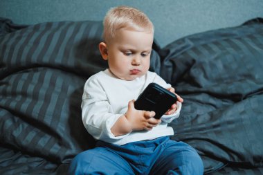 Neşeli küçük çocuk bütün gün yatakta oturup akıllı telefondan oyun oynuyor. Evde akıllı telefon tutarak yatağında oturan çocuğu yakından çek. Cep telefonuyla gülümseyen çocuk.