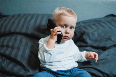 Neşeli küçük çocuk bütün gün yatakta oturup akıllı telefondan oyun oynuyor. Evde akıllı telefon tutarak yatağında oturan çocuğu yakından çek. Cep telefonuyla gülümseyen çocuk.