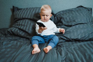 2 yaşında akıllı telefon kullanan, ekrana bakan, elinde telefon tutan, koltukta tek başına oturan, mobil cihazda oyun oynayan, çevrimiçi çizgi film izleyen tatlı bir çocuk.