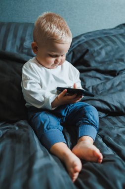 2 yaşında akıllı telefon kullanan, ekrana bakan, elinde telefon tutan, koltukta tek başına oturan, mobil cihazda oyun oynayan, çevrimiçi çizgi film izleyen tatlı bir çocuk.