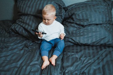 2 yaşında akıllı telefon kullanan, ekrana bakan, elinde telefon tutan, koltukta tek başına oturan, mobil cihazda oyun oynayan, çevrimiçi çizgi film izleyen tatlı bir çocuk.