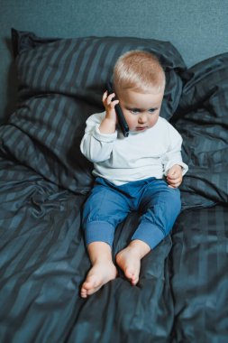 Akıllı telefonu olan 2 yaşında bir çocuk. Küçük çocuk yatakta oturuyor ve cep telefonuyla oynuyor. Annemi arıyorum. Elinde cep telefonu olan sevimli bir çocuk ekranda dikkatle bakıyor..